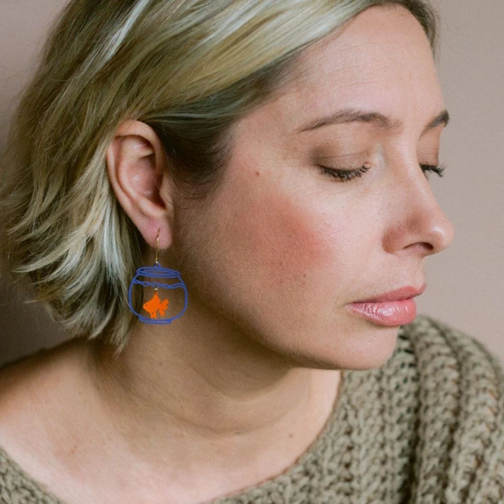 A woman with short blonde hair and closed eyes wears Denz Mini Fish Bowls Orange/Blue Earrings, featuring a blue bowl with an orange fish. She is dressed in a textured, olive-green sweater.