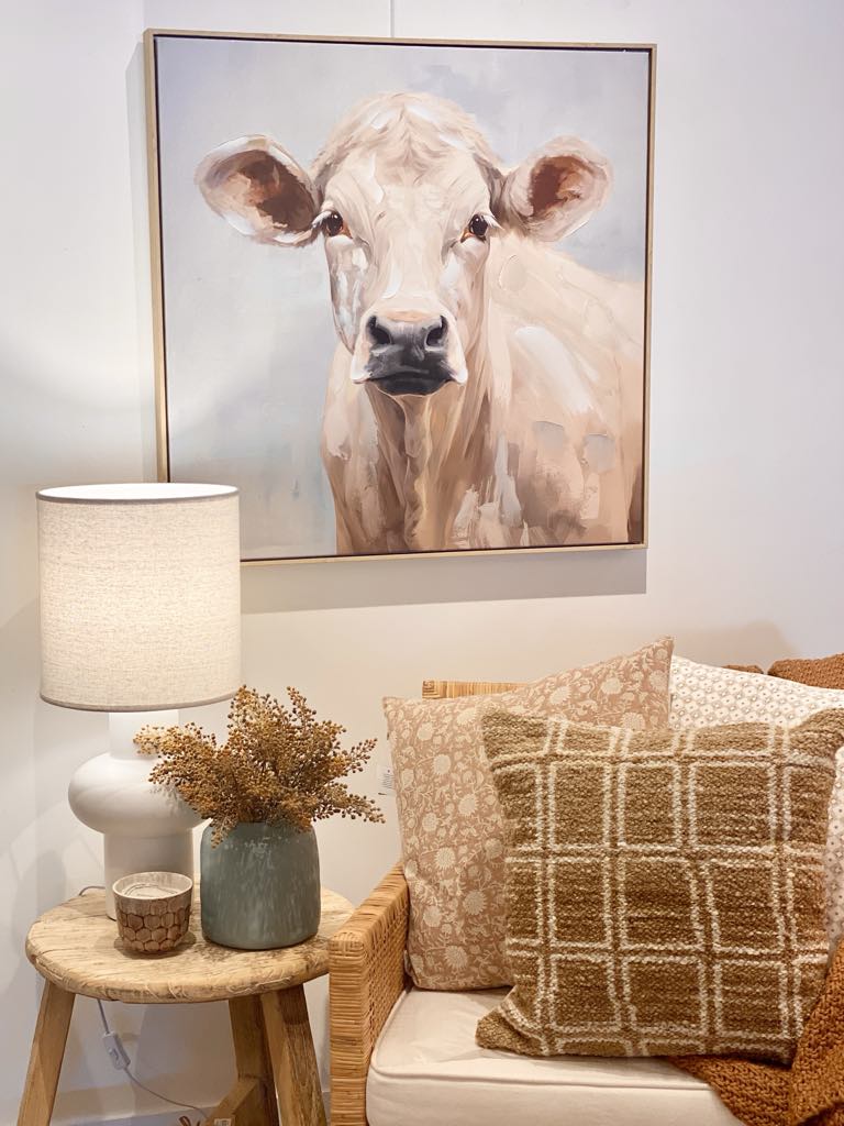 A cozy living room corner with a beige sofa, patterned pillows, a wooden side table with a lamp and vase of dried flowers, and the NF Living Cow Print 83 x 83cm hanging on the wall above.